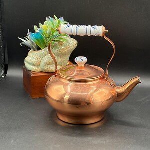 May include: A polished copper tea kettle with a rounded body and a short spout. The kettle features a white ceramic handle and a matching lid knob, both decorated with blue painted leaf patterns. The metal surface reflects light clearly.
