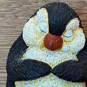 May include: A small figurine of a penguin with a textured black and white finish. The penguin has its eyes closed, a reddish-brown beak, and its wings are folded against its chest. The surface of the figurine has a rough, tactile appearance.