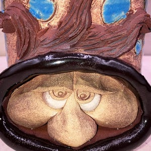 May include: A ceramic mug with a whimsical face, featuring a large nose, wide eyes, and a black rim. The mug is decorated with brown and blue glaze, creating a unique and artistic design.