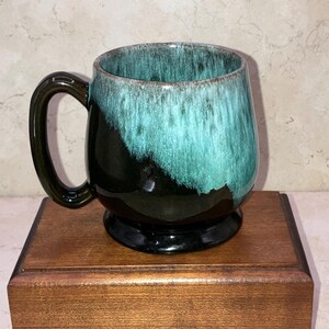 May include: A ceramic mug with a dark green handle and base. The mug's body transitions from a dark green to a turquoise color. The mug is sitting on a wooden box.