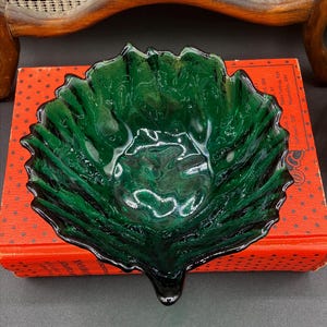 May include: A dark green glass bowl with a leaf-like design. The bowl has a ruffled edge and sits on a red box. The bowl's color is a deep emerald green with black accents, creating a striking contrast.