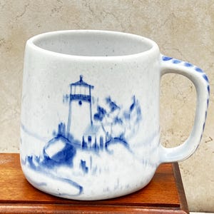May include: White ceramic mug with a blue lighthouse design. The mug features a cylindrical shape with a rounded handle adorned with blue dots. The lighthouse scene is painted in blue, set against a white background. The mug is approximately 4 inches tall.