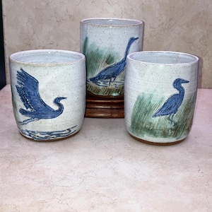 May include: Three ceramic cups with a speckled, light blue glaze. Each cup features a hand-painted blue heron design. The heron is depicted in different poses, with one in flight and others standing in water or grass. The cups are cylindrical and appear handmade.