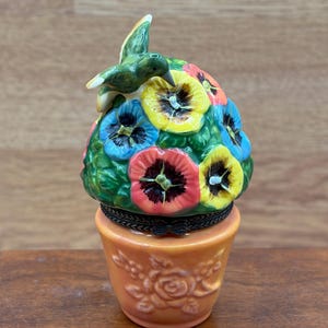 May include: A porcelain trinket box shaped like a flower pot with a lid featuring a cluster of colorful pansies and a green hummingbird perched on top. The orange base is decorated with embossed floral patterns and a dark metal hinge closure.