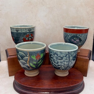 May include: Four ceramic teacups with intricate blue and red floral designs. The cups are displayed on a dark wooden stand, showcasing the detailed patterns and colors. The teacups have a traditional Japanese aesthetic.
