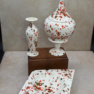 May include: A set of white ceramic decorative items with red and green splatter paint. The set includes a vase, a lidded jar, and an ashtray. The ashtray is square with a rounded interior. The jar is on a pedestal base.