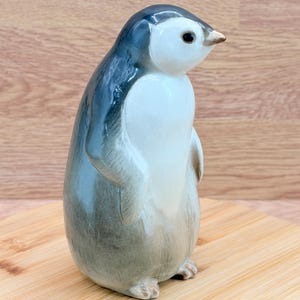 May include: A ceramic penguin figurine featuring a glossy finish with a white chest and face, contrasted by dark gray and blue-toned feathers on the back and sides. The penguin has a small, light-colored beak and dark, round eyes, standing upright.