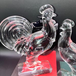 May include: Two clear glass rooster figurines. The rooster on the left has a detailed tail and stands on a rectangular base. The rooster on the right is facing the left and has a simpler design.