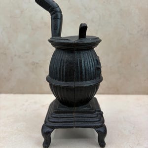 May include: A small, black cast iron stove with a chimney and a square base. The stove has a rounded body with vertical lines and sits on four curved legs. The chimney curves upwards and to the left.