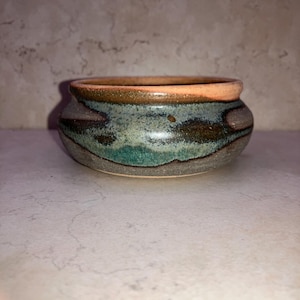 May include: A small, handcrafted ceramic bowl with a rounded shape. The bowl features a color palette of teal, brown, and gray, with a reddish-orange rim. The exterior has a textured, glazed finish, suggesting a rustic or artisanal aesthetic. This decorative bowl is suitable for small items.