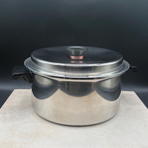 May include: A stainless steel cooking pot with a matching lid. The pot features two black side handles and a central black knob on the lid with a copper-colored accent ring. The exterior surface is polished and reflective, showing a cylindrical shape.