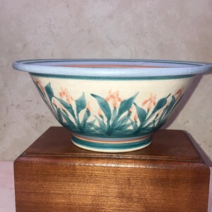 May include: A ceramic bowl with a light blue rim and a floral design. The bowl features a cream-colored base with teal and orange flowers and green leaves. The bowl is sitting on a wooden stand.