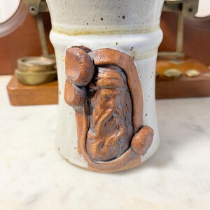 May include: A cylindrical ceramic mug with a speckled off-white glaze and a smooth finish. The front features a raised, dark brown clay relief sculpture of a bearded figure wearing a hat, integrated into the body of the vessel.