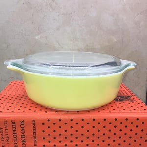 May include: A vintage, oval-shaped, yellow Pyrex casserole dish with a clear glass lid. The dish has two handles and is sitting on a red book with black polka dots. The dish is a pale yellow color.