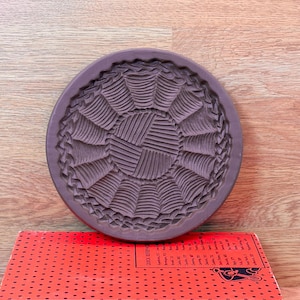 May include: A circular, dark brown ceramic mold featuring a detailed, repeating radial pattern of textured, fan-like segments and a central circular motif with fine, etched lines, resting atop a bright red book with black polka dots.