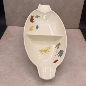 May include: Cream-colored, oval-shaped serving dish with two compartments and a handle. The dish is decorated with colorful autumn leaves in shades of red, yellow, blue, and green. A vintage piece for serving snacks or appetizers.