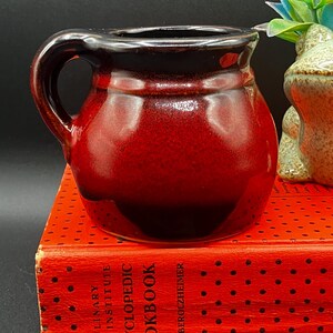 May include: A small, handcrafted ceramic mug with a dark red glaze that fades to black at the rim. The mug has a rounded body, a sturdy handle, and sits on a red book. The book's spine reads "CULINARY ARTS INSTITUTE ENCYCLOPEDIC COOKBOOK".