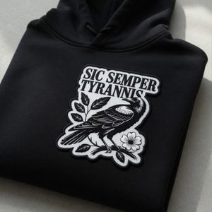 Puede incluir: Sudadera con capucha negra con un parche que muestra una ilustración de un cuervo y el texto "SIC SEMPER TYRANNIS". El cuervo está representado en blanco y negro, posado en una rama con una flor.