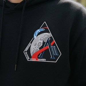 Puede incluir: Sudadera con capucha negra con un parche triangular que representa una luna y un cohete. El parche incluye los nombres "WISEMAN", "GLOVER", "KOCH" y "HANSEN". El diseño incorpora acentos rojos, azules y blancos.