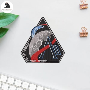 Puede incluir: Un parche triangular negro con una luna estilizada y la letra "A" con detalles rojos y blancos. El parche incluye los nombres "WISEMAN", "GLOVER", "KOCH" y "HANSEN". Probablemente un emblema de astronauta o misión espacial.