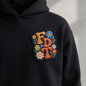Puede incluir: Sudadera con capucha negra con un parche bordado. El parche presenta las letras "FDT" en naranja, rodeadas de un diseño floral con flores azules, amarillas y rosas. La sudadera está hecha de un material suave.