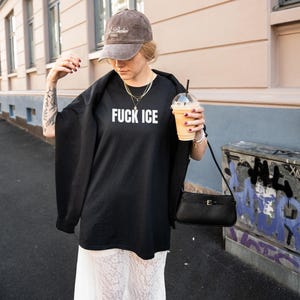 K&ouml;nnte beinhalten: Schwarzes &uuml;bergro&szlig;es T-Shirt mit dem wei&szlig;en Text "FUCK ICE", kombiniert mit einer braunen Kappe und einer schwarzen Handtasche. Die Person h&auml;lt ein Getr&auml;nk in einem transparenten Becher mit Strohhalm.