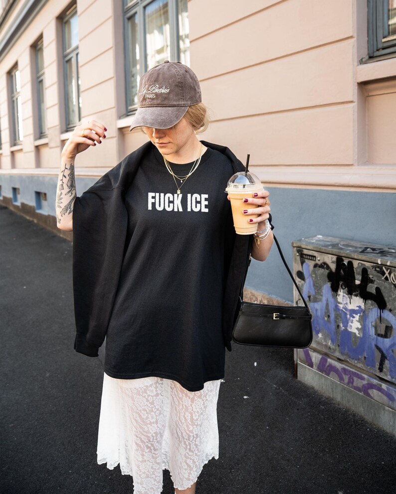 K&ouml;nnte beinhalten: Schwarzes &uuml;bergro&szlig;es T-Shirt mit dem wei&szlig;en Text "FUCK ICE", kombiniert mit einer braunen Kappe und einer schwarzen Handtasche. Die Person h&auml;lt ein Getr&auml;nk in einem transparenten Becher mit Strohhalm.