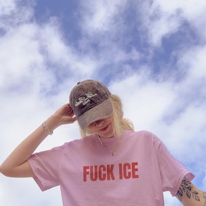 K&ouml;nnte beinhalten: Hellrosa T-Shirt mit dem Schriftzug "FUCK ICE" in roten Blockbuchstaben. Die Person tr&auml;gt eine braune Baseballkappe mit wei&szlig;em Text und eine goldene Halskette. Der Hintergrund ist ein blauer Himmel mit wei&szlig;en Wolken.