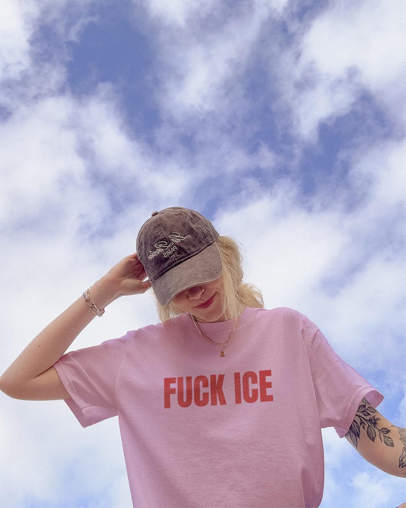 K&ouml;nnte beinhalten: Hellrosa T-Shirt mit dem Schriftzug "FUCK ICE" in roten Blockbuchstaben. Die Person tr&auml;gt eine braune Baseballkappe mit wei&szlig;em Text und eine goldene Halskette. Der Hintergrund ist ein blauer Himmel mit wei&szlig;en Wolken.