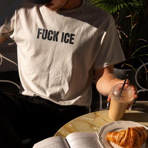 K&ouml;nnte beinhalten: Beiges T-Shirt mit dem Text "FUCK ICE" in fetten schwarzen Buchstaben. Das Bild zeigt auch ein Croissant auf einem Teller, ein Kaffeegetr&auml;nk und ein offenes Buch auf einem Tisch.