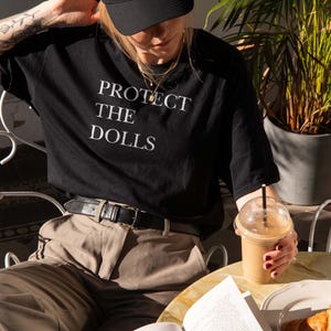 May include: A person wearing a black t-shirt with the text "PROTECT THE DOLLS" in white. They are also wearing a black cap and tan pants. A drink, book, and croissant are on the table.