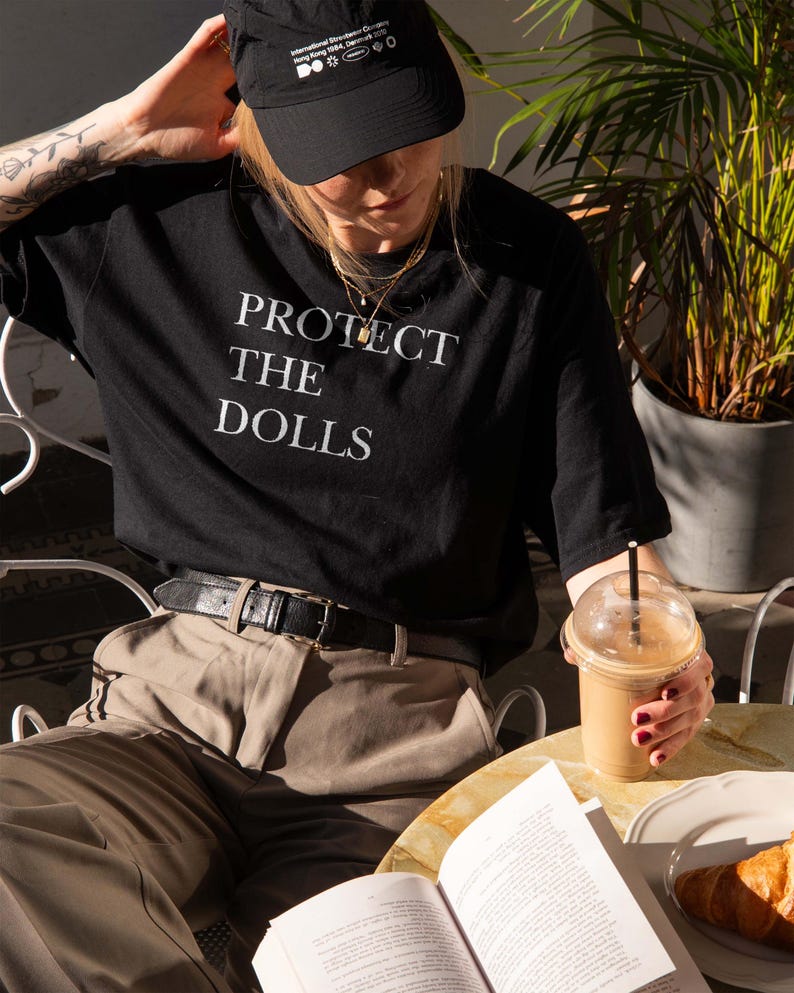 May include: A person wearing a black t-shirt with the text "PROTECT THE DOLLS" in white. They are also wearing a black cap and tan pants. A drink, book, and croissant are on the table.