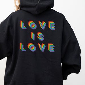 Könnte beinhalten: Schwarzer Kapuzenpullover mit dem Schriftzug "LOVE IS LOVE" in regenbogenfarbenen 3D-Buchstaben. Jeder Buchstabe enthält einen Regenbogenbogen, wodurch ein lebendiges und auffälliges Design entsteht. Ein Statement-Kleidungsstück.
