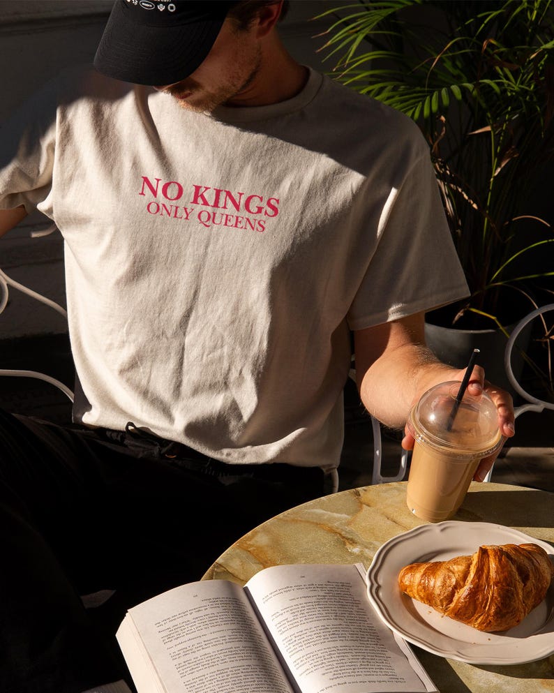 Op de afbeelding: Een beige t-shirt met de roze tekst "NO KINGS ONLY QUEENS". De afbeelding toont een persoon die een koffiedrank met een rietje vasthoudt, een croissant op een bord en een open boek op een tafel.