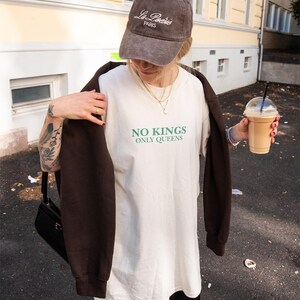 Op de afbeelding: Bruine baseballcap met geborduurd "La Po&eacute;sie Paris". De persoon draagt een wit t-shirt met de groene tekst "NO KINGS ONLY QUEENS". Een bruin sweatshirt is over de schouders gedrapeerd. De persoon houdt een drankje vast.