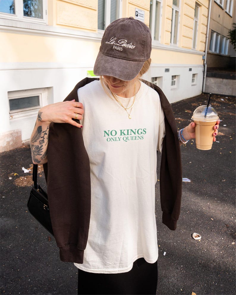 Op de afbeelding: Bruine baseballcap met geborduurd "La Po&eacute;sie Paris". De persoon draagt een wit t-shirt met de groene tekst "NO KINGS ONLY QUEENS". Een bruin sweatshirt is over de schouders gedrapeerd. De persoon houdt een drankje vast.