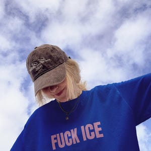 K&ouml;nnte beinhalten: Ein blaues Sweatshirt mit dem Text "FUCK ICE" in rosa Buchstaben. Die Person tr&auml;gt eine braune Kappe mit wei&szlig;em Text. Der Hintergrund ist ein blauer Himmel mit wei&szlig;en Wolken.