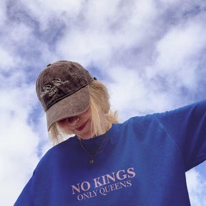Op de afbeelding: Een blauwe trui met de tekst "NO KINGS ONLY QUEENS" in roze letters. Een bruine baseballpet met witte tekst wordt gedragen. De achtergrond is een bewolkte lucht.