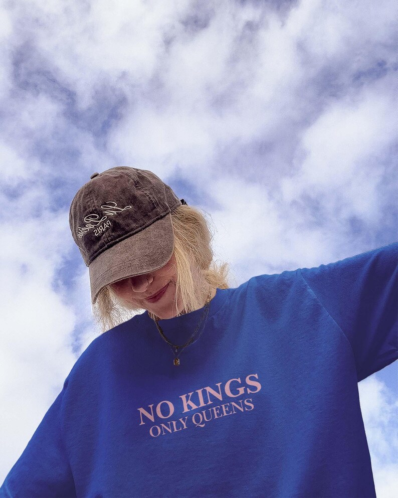 Op de afbeelding: Een blauwe trui met de tekst "NO KINGS ONLY QUEENS" in roze letters. Een bruine baseballpet met witte tekst wordt gedragen. De achtergrond is een bewolkte lucht.