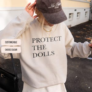 May include: A beige sweatshirt with the text "PROTECT THE DOLLS" in black. The image also shows a brown cap with "La Poche PARIS" written on it. Two buttons on the left say "CUSTOMIZABLE" and "CHOOSE COLOUR".