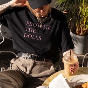 May include: Black t-shirt with the text "PROTECT THE DOLLS" in pink. The person is wearing a black cap and tan pants. A cup of coffee and a book are on the table.
