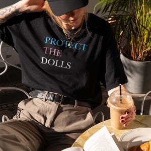 May include: Black t-shirt with the text "PROTECT THE DOLLS" in blue, pink, and white. The person is wearing a black cap and tan pants. A cup of coffee and a croissant are on the table.