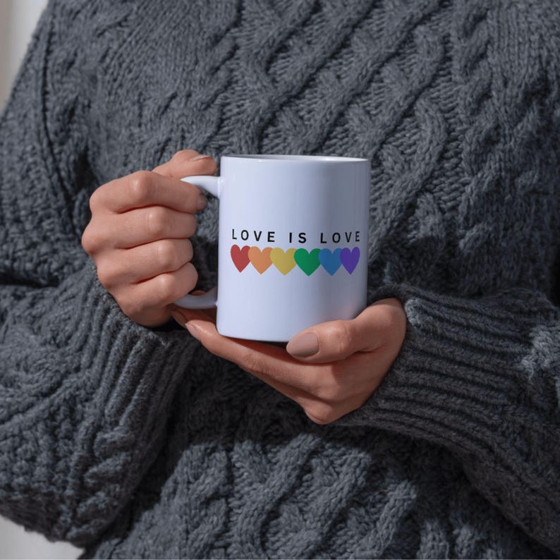 May include: White ceramic mug with the text "LOVE IS LOVE" and a row of rainbow-colored hearts. The mug is held by a person wearing a gray cable-knit sweater. The mug is a standard 11 oz size.
