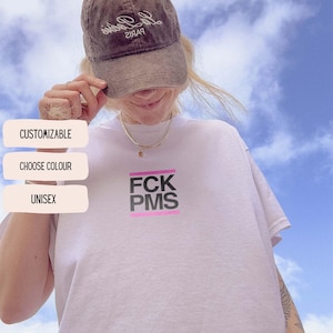 May include: White unisex t-shirt with the text "FCK PMS" in black and pink. The image also shows a brown baseball cap and the words "Customisable", "Choose Colour", and "Unisex" in a white box.
