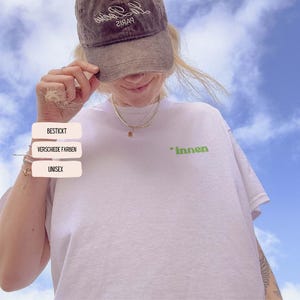 Pode incluir: Uma camiseta branca com a palavra "innen" em verde. A imagem também mostra um boné marrom com texto e três caixas de texto que dizem "BESTICKT", "VERSCHIEDE FARBEN" e "UNISEX".