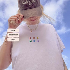 Peut inclure: T-shirt blanc avec les lettres brodées "FCK MRZ" aux couleurs de l'arc-en-ciel. Le t-shirt est unisexe et porte les mots "BESTICKT", "VERSCHIEDE FARBEN" et "UNISEX" sur le côté gauche. Une casquette marron est portée.
