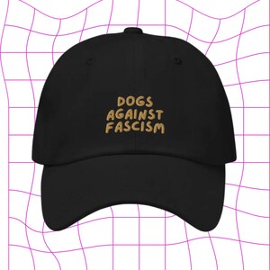 Może przedstawiać: Czarna czapka z daszkiem z haftem "DOGS AGAINST FASCISM" w kolorze złotym z przodu.