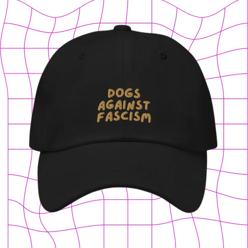 Może przedstawiać: Czarna czapka z daszkiem z haftem "DOGS AGAINST FASCISM" w kolorze złotym z przodu.