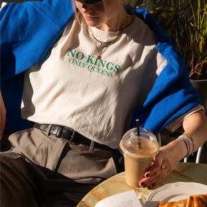 Op de afbeelding: Een persoon draagt een beige t-shirt met de groene tekst "NO KINGS ONLY QUEENS". De persoon draagt een bruine pet en een blauwe trui. Een zwarte riem, een kopje koffie, een croissant en een open boek zijn ook zichtbaar.