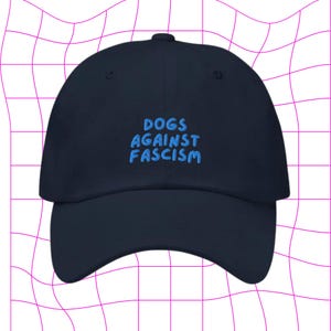 Może przedstawiać: Czapka bejsbolowa w kolorze granatowym z haftem "DOGS AGAINST FASCISM" w jasnoniebieskim kolorze.